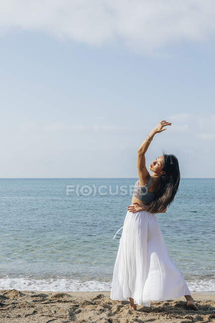 Обратный вид изящной молодой этнической женщины, делающей обратный сгиб с поднятой рукой, практикующей йогу на песчаном побережье против океана — стоковое фото