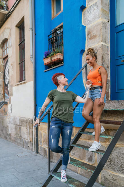 Cool smiling young homosexual girlfriends holding hands walking down staircase against building in town — Stock Photo