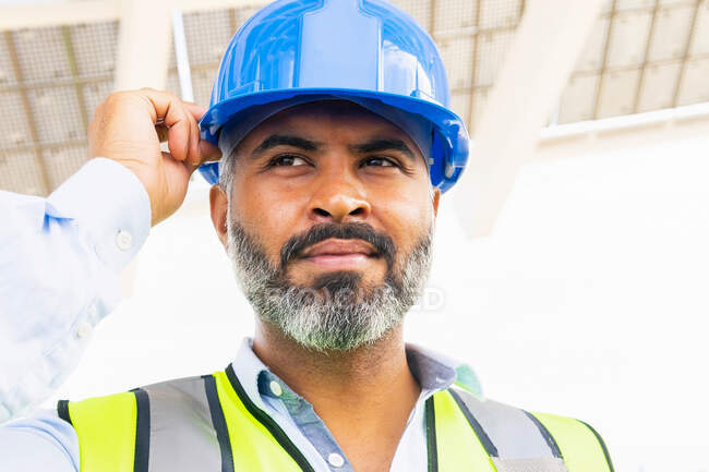 Mature ethnic male supervisor in vest putting on hardhat while looking away standing near solar power station — Stock Photo