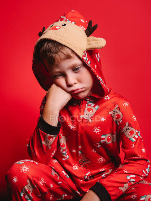 Bored cute little boy in hooded pajama with deer leaning on hand and looking down while sitting on haunches in red studio — Stock Photo