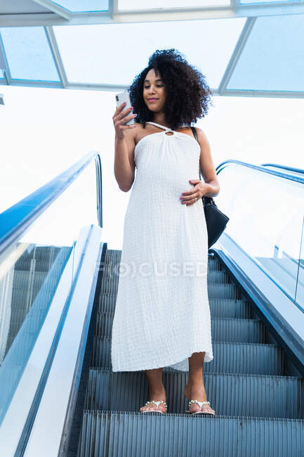 Desde abajo mujer afroamericana anticipando bebé de pie en escaleras móviles y navegando por el teléfono celular - foto de stock