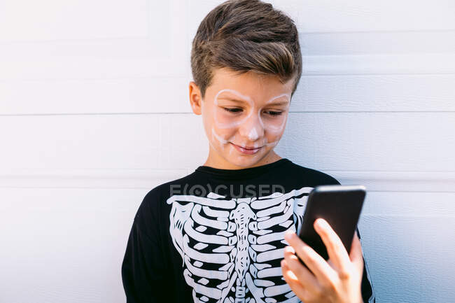 Niño alegre en traje de esqueleto negro con la cara pintada usando en el teléfono móvil mientras está sentado cerca de la pared blanca en la calle - foto de stock