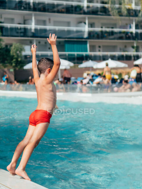 Ganzkörper-Rückenansicht eines hemdslosen Jungen, der die Hände hebt, während er vom Beckenrand abstößt und ins Wasser springt — Stockfoto