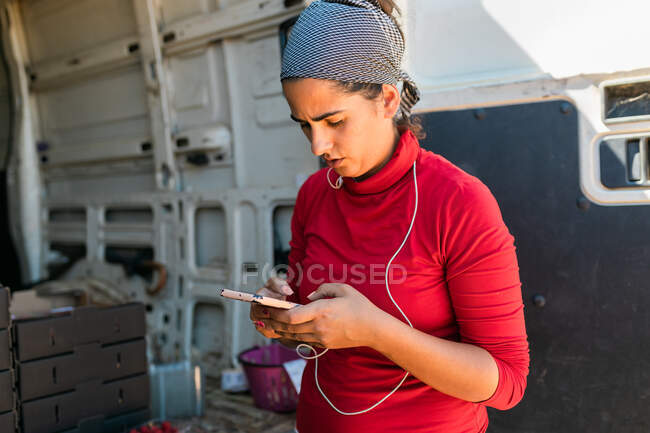 Jardinero femenino concentrado en bandana que navega por el teléfono celular mientras trabaja en la granja y mira la pantalla - foto de stock