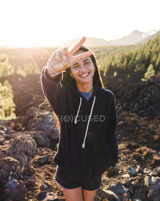 Mujer franca adolescente en sudadera demostrando gesto de victoria mientras mira a la cámara contra el monte en Tenerife con la espalda iluminada - foto de stock