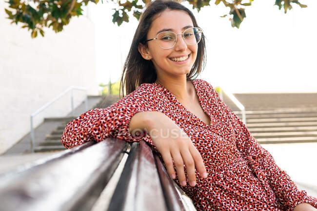 Mujer joven positiva en ropa elegante sentado mirando a la cámara en el banco de madera en el parque - foto de stock