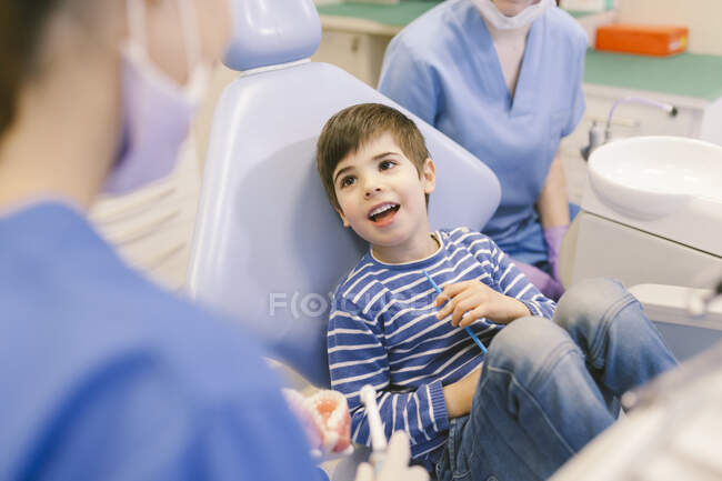 Высокий угол у мальчика с открытым ртом, разговаривающего с врачами во время стоматологического лечения в кабинете современного стоматолога — стоковое фото