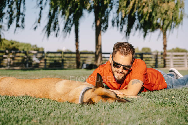 Уровень грунта молодого бородатого мужчины в солнечных очках, лежащего на лугу против чистокровной собаки в парке в летний день — стоковое фото