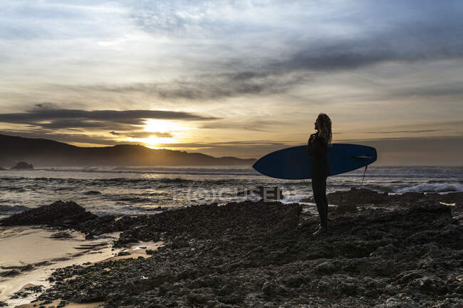 Вид сбоку на неузнаваемую молодую женщину, стоящую на берегу с доской для серфинга, прежде чем попасть в море во время заката на пляже в Астурии, Испания — стоковое фото