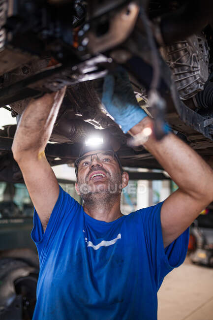 Busy mature male mechanic in head torch repairing automobile placed on elevator in workshop — Stock Photo