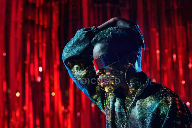 Jeune femme noire branchée dans des lunettes cyberpunk avec des ombres sur le visage dans des rayons de lumière dans une boîte de nuit — Photo de stock
