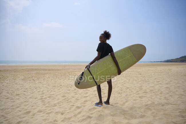 Вид афроамериканской спортсменки с доской для серфинга, любующейся океаном с песчаного берега под облачным голубым небом — стоковое фото