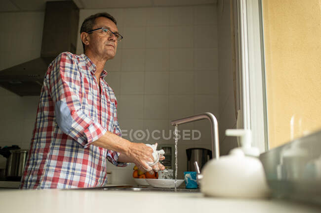 Vista lateral da calma pensativo maduro macho lavar pratos sujos enquanto está perto de pia na cozinha e fazer tarefas domésticas — Fotografia de Stock