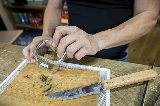 Cultivez un mâle méconnaissable avec un couteau coupant une plante de cannabis séchée sur une planche de bois dans un espace de travail — Photo de stock