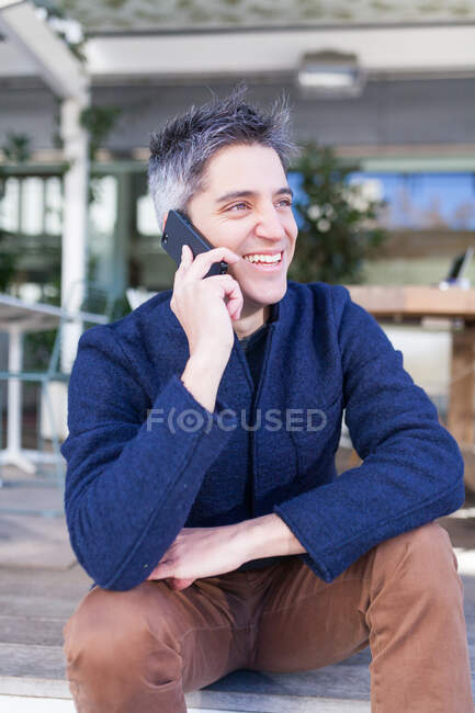 Щасливий молодий чоловік в повсякденному одязі посміхається, сидячи на лавці і відповідаючи на телефонний дзвінок — стокове фото