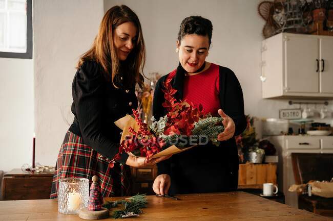 Alegres amigas de pie a la mesa con velas y haciendo creativos ramos de Navidad para la celebración de las fiestas - foto de stock