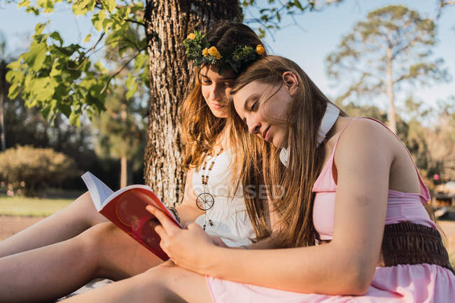 Novias en vestidos de novia compartiendo libro de texto mientras están sentadas en el prado en el soleado parque en la parte posterior iluminada - foto de stock