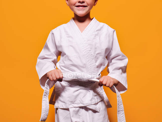 Anonyme sourire garçon en kimono blanc serrant noeud ceinture de karaté avec ses mains sur fond orange vif et en regardant la caméra — Photo de stock