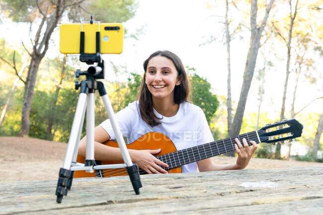 Cheerful female teen blogger playing acoustic guitar while recording video on cellphone on tripod in park — Stock Photo