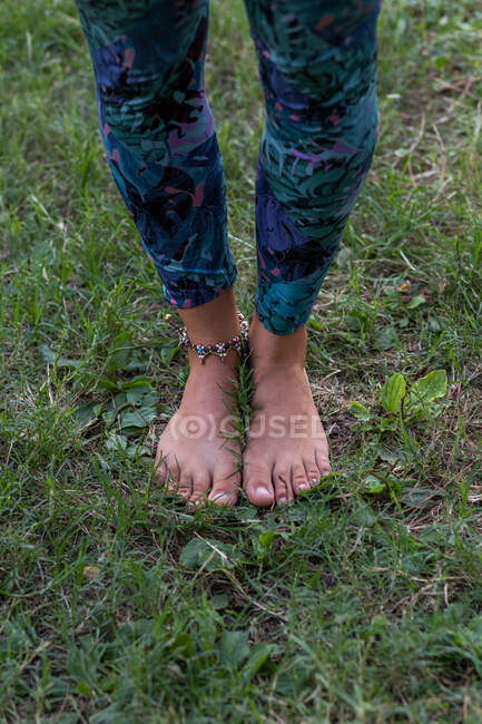 De arriba de la cosecha irreconocible hembra descalza en leggins de pie sobre hierba verde en la naturaleza a la luz del día - foto de stock
