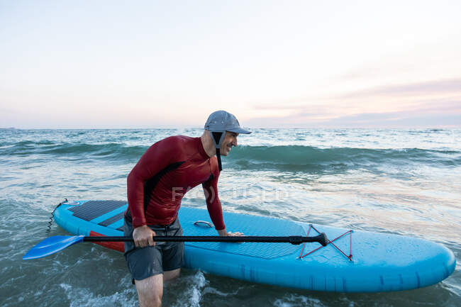 Vista lateral do surfista masculino em fato de mergulho e chapéu carregando prancha de remo e entrando na água para surfar na praia — Fotografia de Stock