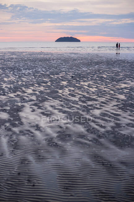 Coppia in piedi sulla riva sabbiosa bagnata da acque poco profonde del mare con collina all'orizzonte sotto il cielo del tramonto in Malesia — Foto stock