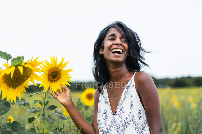 Sincero adulto fêmea étnica olhando para longe no prado tocando flores florescentes no campo no fundo borrado — Fotografia de Stock