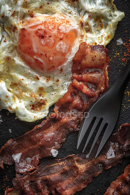 De cima do lado ensolarado para cima ovo com fatias de bacon frito e condimentos na bandeja contra talheres no fundo escuro — Fotografia de Stock