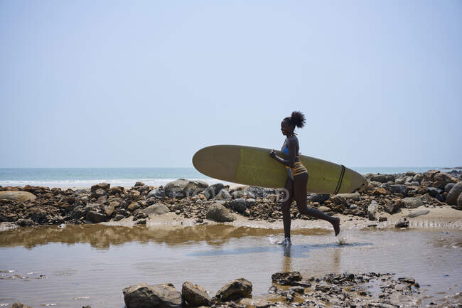 Side view of young ethnic sportswoman in swimsuit with Afro hair bun and surfboard looking away while running on stone coast — Stock Photo