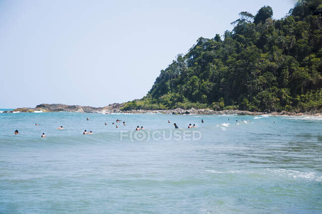 Scenic view of tourists swimming in stormy sea against mountain with lush green trees under light sky — Stock Photo