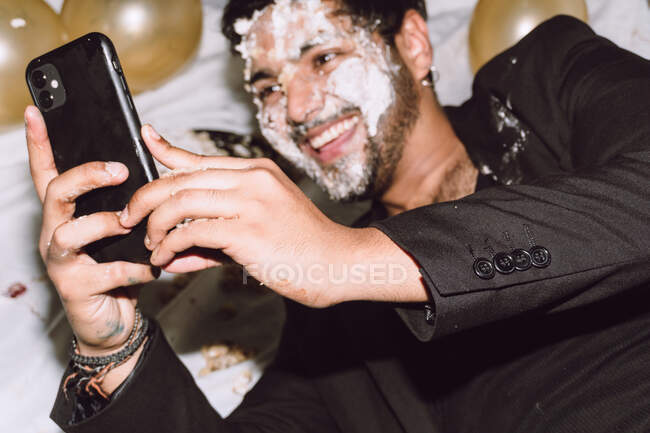 Врожай бородатий позитивний чоловік з розбитим тортом на обличчі, лежить серед кульок і приймає власний портрет на мобільний телефон під час вечірки на день народження — стокове фото