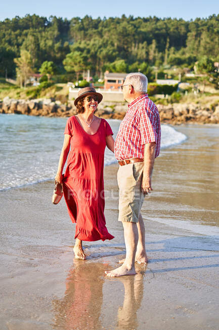 Sonriendo descalza pareja de ancianos en gafas de sol de pie mirándose unos a otros en la playa de arena húmeda y disfrutando de un día soleado - foto de stock
