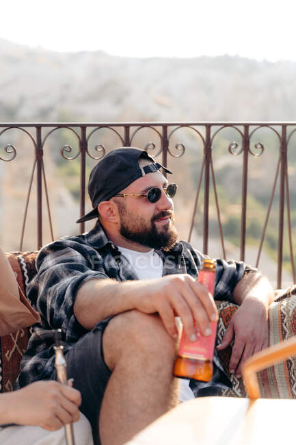 Взрослый бородатый мужчина в стильных солнцезащитных очках сидит на диване с бутылкой пива рядом с другом урожая — стоковое фото