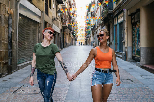 Круті молоді гомосексуальні жінки з татуюваннями в сонцезахисних окулярах тримають руки на прогулянці в місті — стокове фото