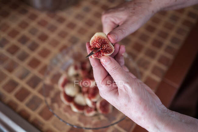 From above of crop unrecognizable elderly person with knife cutting ripe fig above bowl on table in house — Stock Photo