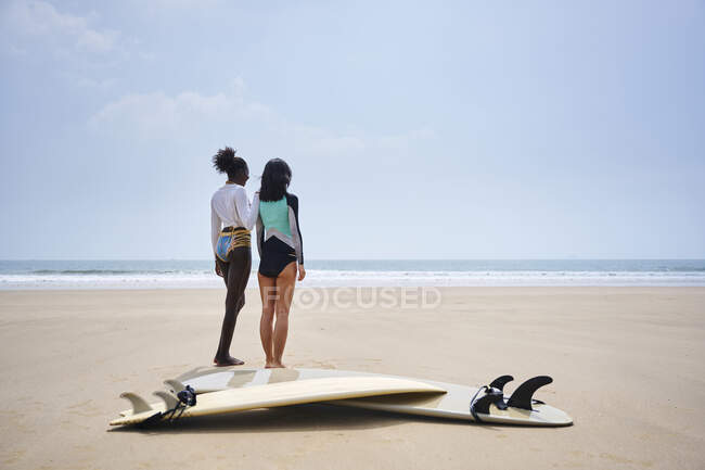 Неузнаваемый серфер во время общения с веселой черной подружкой против доски для серфинга и серфинга на песчаном побережье — стоковое фото