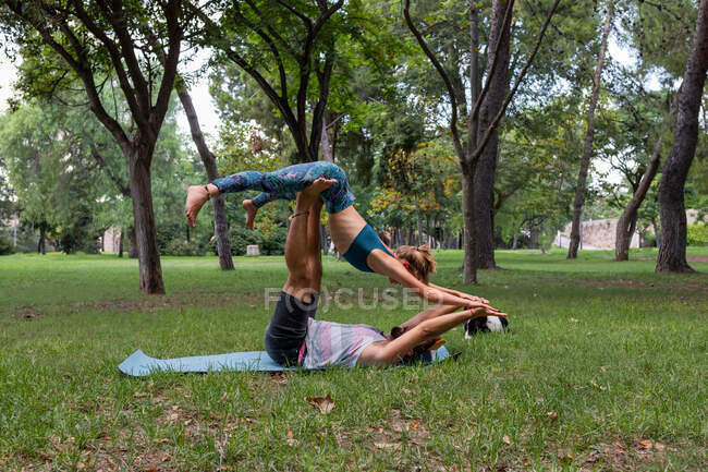 Вид сбоку гибкой пары, которая в дневное время вместе занимается акройогой на коврике для йоги на траве против деревьев в парке — стоковое фото