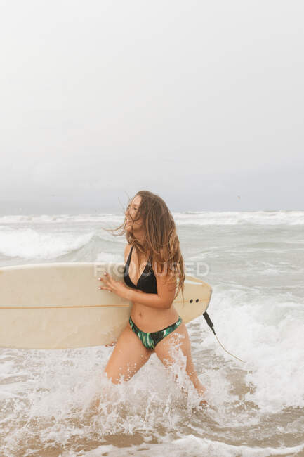 Side view of young cheerful sportswoman with flying hair and surfboard in ocean with foam under cloudy sky — Stock Photo