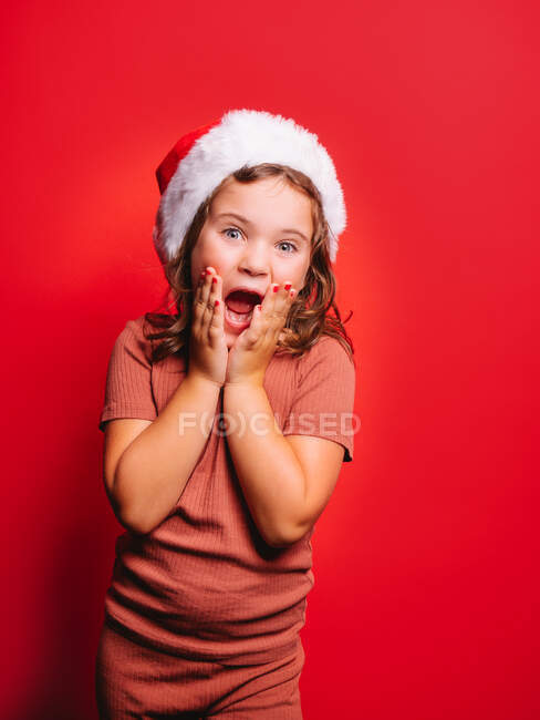 Menina engraçada surpreendida em roupas casuais e chapéu de Papai Noel com cabelo ondulado e manicure tocando bochechas e gritando contra fundo vermelho — Fotografia de Stock