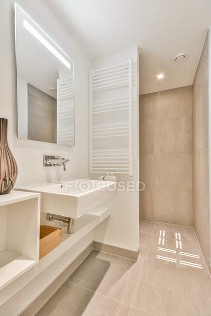 Moderno interno del bagno con lavabo in ceramica bianca sotto specchio illuminato e pavimento e pareti piastrellate beige — Foto stock