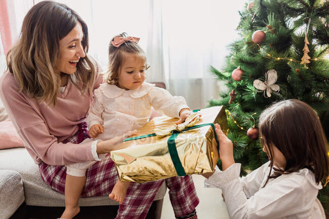 Fröhliche Mutter mit Kleinkind-Tochter übergibt Geschenkbox an Mädchen gegen geschmückten Tannenbaum beim Neujahrsfest im Haus — Stockfoto
