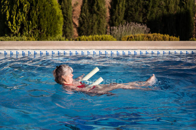 Вид сбоку жизнерадостной пожилой женщины с длинными волосами, плавающей в бассейне с аквалапшой — стоковое фото