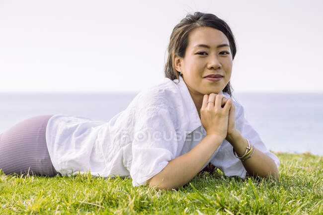 Сексуальна молода етнічна жінка в повсякденному одязі дивиться на камеру, лежачи на зеленому узбережжі вдень — стокове фото