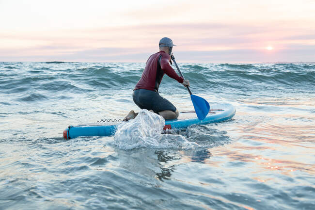Back view of unrecognizable male surfer in wetsuit and hat on paddle board surfing on seashore — Stock Photo