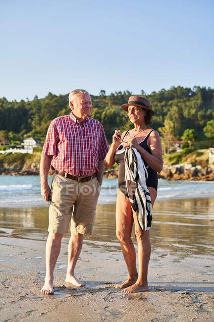 Sorrindo descalço casal de idosos em óculos de sol em pé na praia de areia molhada e desfrutando de dia ensolarado — Fotografia de Stock