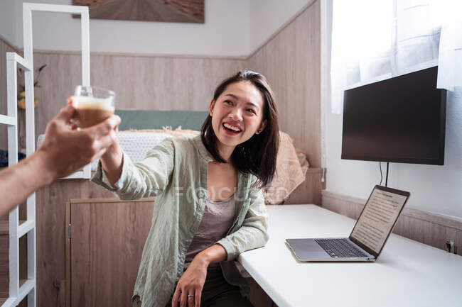 Jovem escritora étnica alegre recebendo copo de café da colheita parceiro anônimo contra laptop em casa — Fotografia de Stock