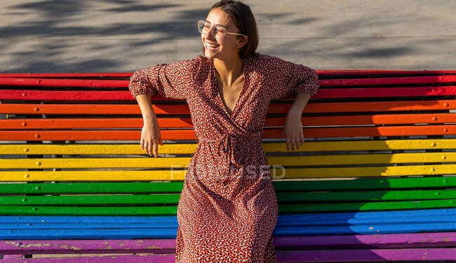 De cima de fêmea alegre em vestido vermelho sentado no banco de madeira colorido e olhando para longe no dia ensolarado — Fotografia de Stock