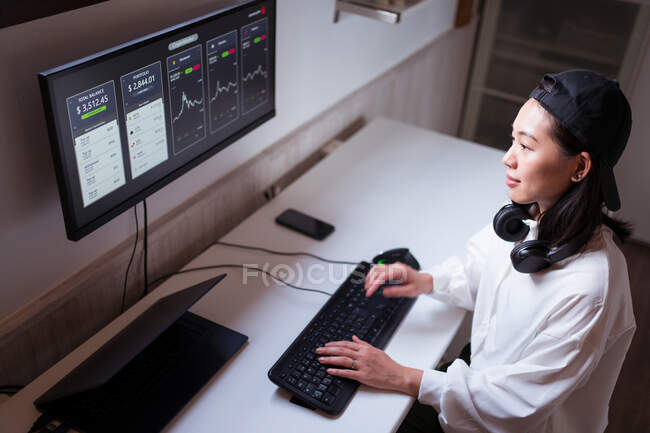 Alto ángulo concentrado mujer asiática que trabaja en la computadora con gráficos que muestran la dinámica de los cambios en el valor de la criptomoneda en el lugar de trabajo conveniente - foto de stock