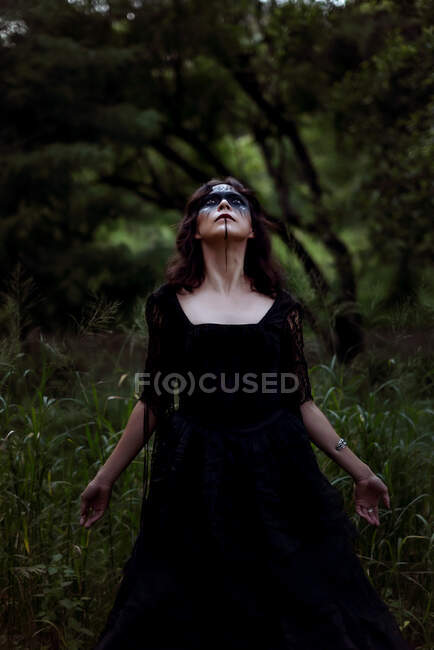 Mystische Hexe im langen schwarzen Kleid und mit aufgemaltem Gesicht, die in dunklen, düsteren Wäldern aufblickt — Stockfoto