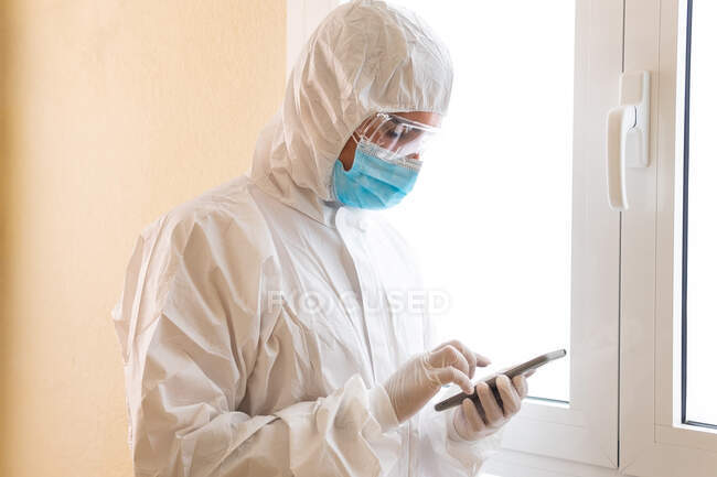 Неузнаваемый мужчина-медик в средствах индивидуальной защиты, касающийся экрана на смартфоне во время пандемии COVID 19 в клинике — стоковое фото